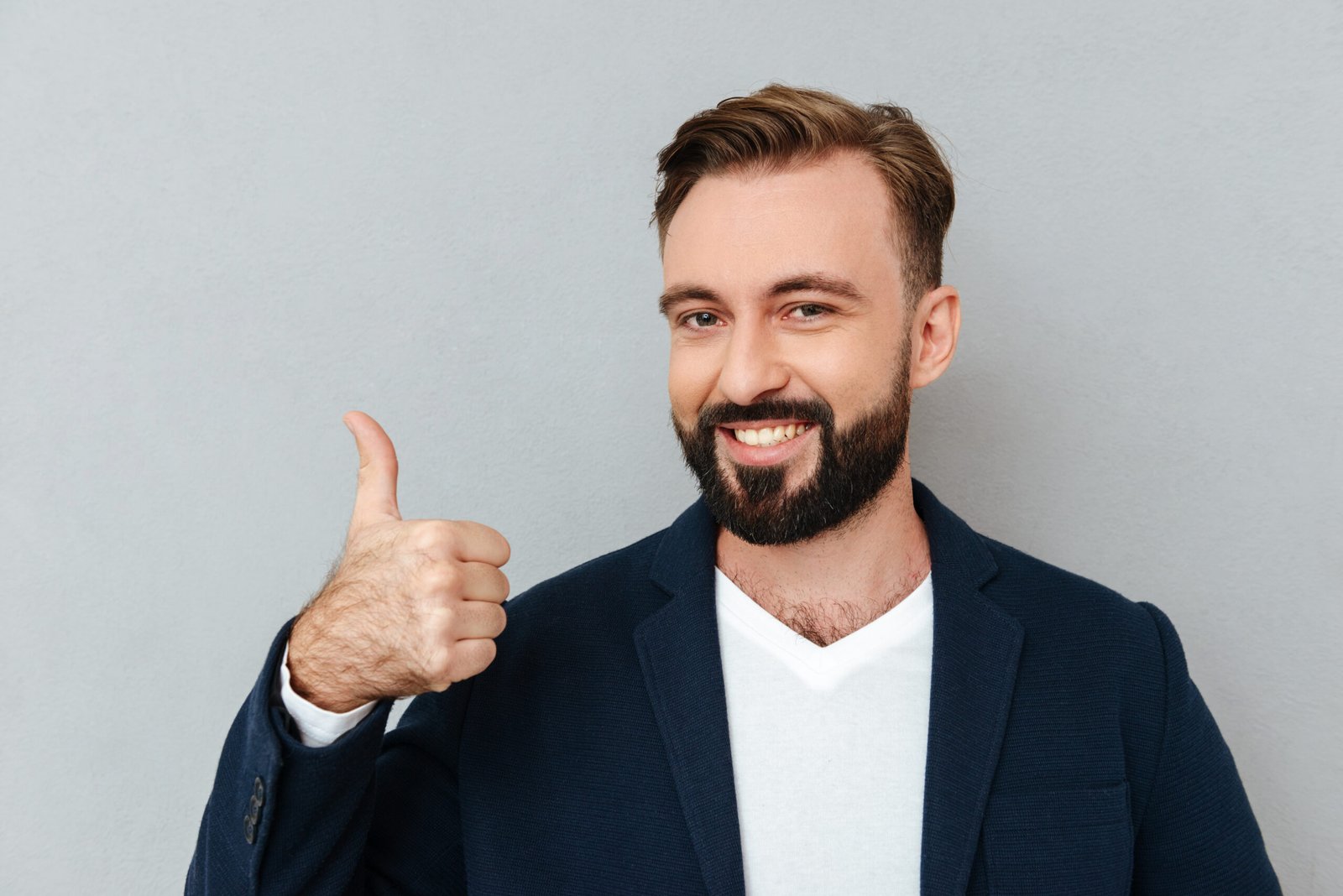 Cheerful bearded man in business clothes showing thumb up and looking at the camera over gray background
