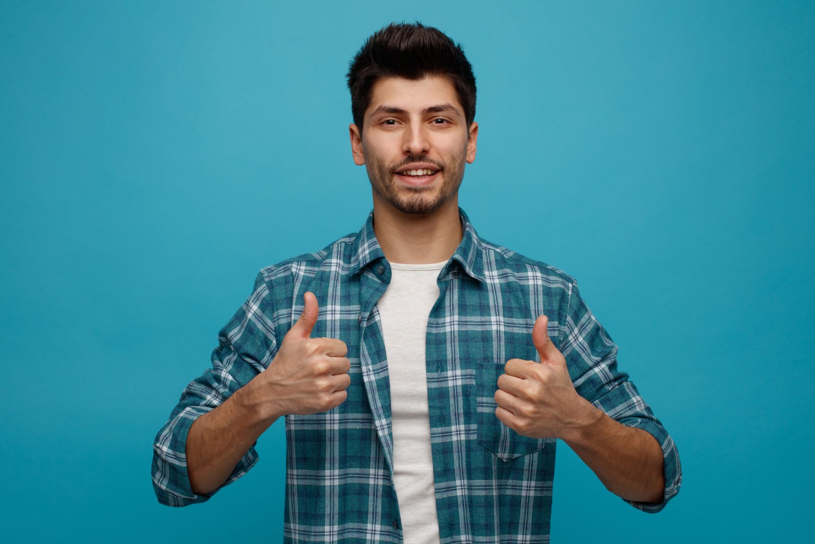 joyful young man looking at camera showing thumbs up isolated on blue background